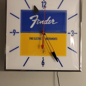 May include: A white clock with black hands and a blue and yellow face. The face has the Fender logo and the words "Fine Electric Instruments".