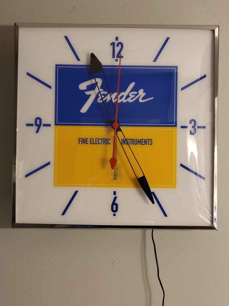May include: A white clock with black hands and a blue and yellow face. The face has the Fender logo and the words "Fine Electric Instruments".