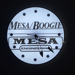 May include: A round, illuminated clock with the text "Mesa/Boogie" and "Mesa Engineering" in white. The clock face is white with black dots marking the hours and black clock hands. The clock is set against a dark background.