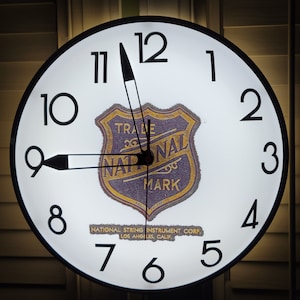 May include: A white clock face with black numbers and hands. The clock has a black border and a gold and purple logo that says "Trade Mark National". The logo is surrounded by the text "National String Instrument Corp. Los Angeles, Calif."