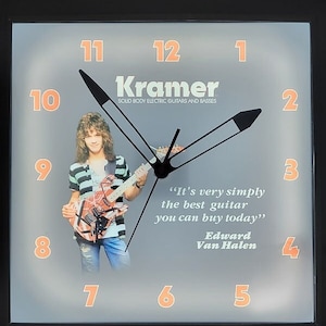 May include: A gray clock with black hands and orange numbers. The clock features a picture of a man playing a red and black electric guitar. The clock also has the text "Kramer" and "SOUD BODY ELECTRIC GUITARS AND BASSES". The quote "It's very simply the best guitar you can buy today" is attributed to "Edward Van Halen".