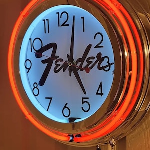 Puede incluir: Un reloj cromado con una esfera blanca y n&uacute;meros negros. El reloj tiene un anillo de ne&oacute;n rojo alrededor del borde y la palabra "Fender" en escritura negra en la esfera.