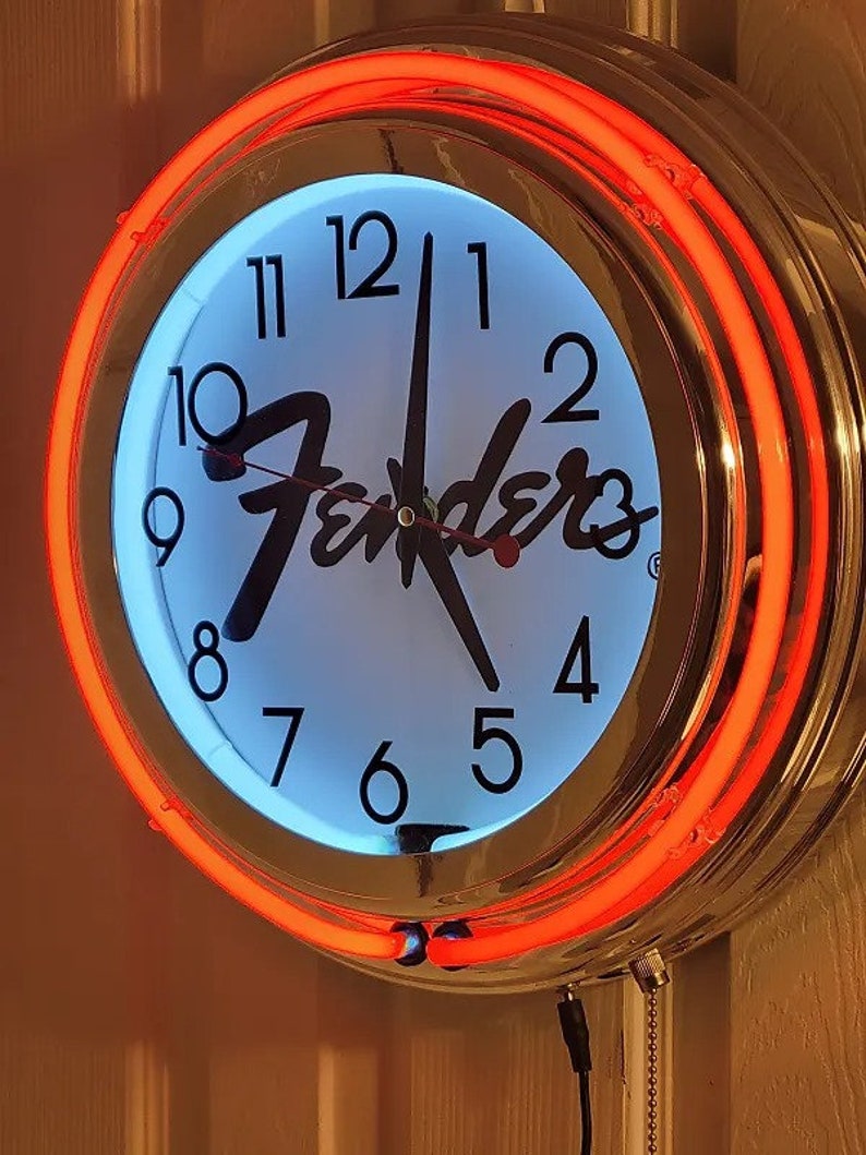 Puede incluir: Un reloj cromado con una esfera blanca y n&uacute;meros negros. El reloj tiene un anillo de ne&oacute;n rojo alrededor del borde y la palabra "Fender" en escritura negra en la esfera.