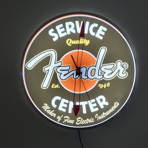 May include: A round, illuminated clock with a brown background and a white border. The clock face features the Fender logo, the words "Service Quality", "Est. 1946", and "Center Maker of Fine Electric Instruments".