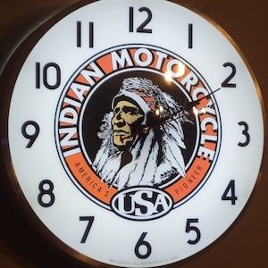 May include: A white wall clock with a black and orange Indian Motorcycle logo. The clock face has black Roman numeral hour markers and black hands. The logo features a Native American man wearing a headdress and the words "Indian Motorcycle" and "America's Pioneer" around the outside. The words "USA" are in a black oval in the center of the logo.