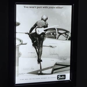 May include: Monochrome framed advertisement showing a person in a flight suit with a guitar on a jet. The text reads "You won't part with yours either." The Fender logo is displayed at the bottom.