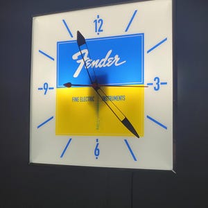 May include: A white square clock with blue and yellow accents. The clock face features the Fender logo and the text "Fine Electric Instruments". The clock hands are black and red.