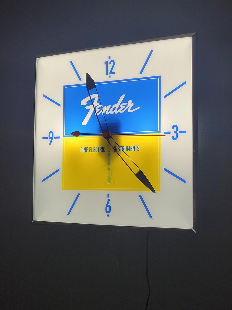 May include: A white square clock with blue and yellow accents. The clock face features the Fender logo and the text "Fine Electric Instruments". The clock hands are black and red.