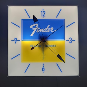May include: A white square clock with blue and yellow accents. The clock face features the Fender logo and the words "Fine Electric Instruments". The clock hands are black and point to the numbers 12 and 6.
