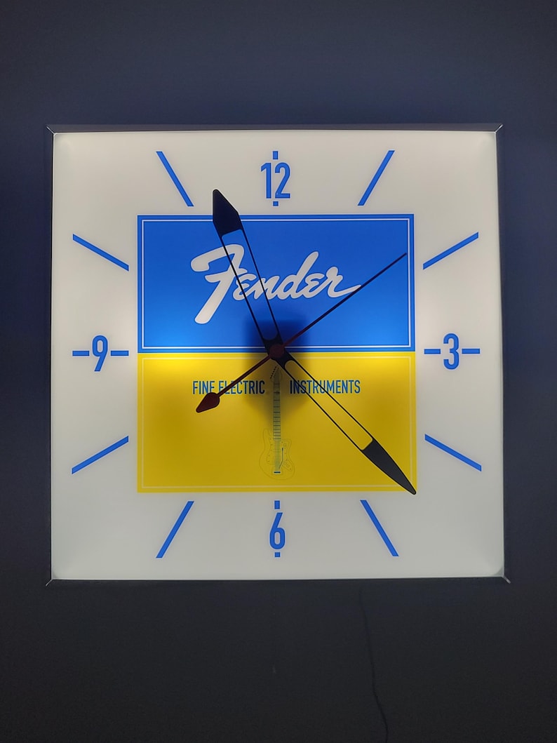 May include: A white square clock with blue and yellow accents. The clock face features the Fender logo and the words "Fine Electric Instruments". The clock hands are black and point to the numbers 12 and 6.