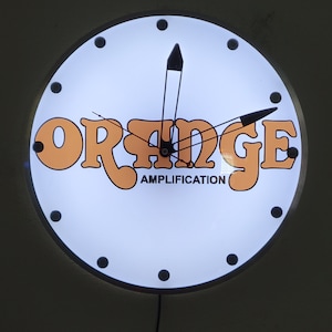 May include: A white round clock with black hands and black dots around the edge. The clock face has the word "ORANGE" in orange letters with the words "AMPLIFICATION" underneath.