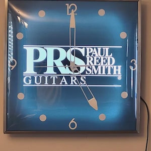 May include: Square wall clock with a blue face and a silver frame. The clock face features the text "PRS PAUL REED SMITH GUITARS" in white and teal. The clock hands point to approximately 4:58.