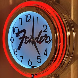 Puede incluir: Un reloj de cromo y ne&oacute;n rojo con una esfera blanca y agujas negras. El reloj presenta el logotipo de Fender y n&uacute;meros negros alrededor del borde.