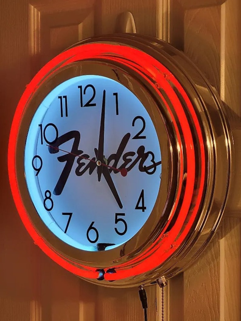 Puede incluir: Un reloj de cromo y ne&oacute;n rojo con una esfera blanca y agujas negras. El reloj presenta el logotipo de Fender y n&uacute;meros negros alrededor del borde.