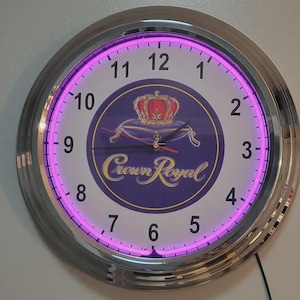 May include: A round, silver-framed wall clock with a purple neon light. The clock face features the Crown Royal logo in a purple and gold design. The numbers are white, and the hands are black.