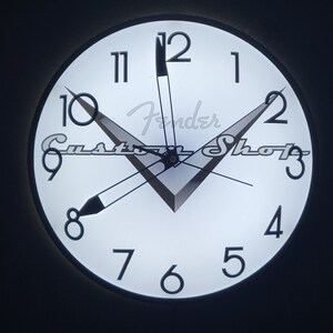May include: A white clock with black numbers and hands. The clock has a Fender Custom Shop logo in the center. The clock is illuminated from behind.