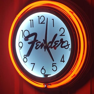 Puede incluir: Una esfera de reloj blanca con n&uacute;meros y manecillas negros, con el logotipo de Fender en negro. El reloj est&aacute; rodeado por un anillo de ne&oacute;n naranja brillante.