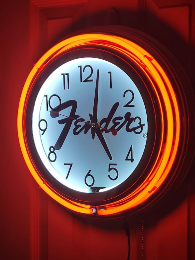 Puede incluir: Una esfera de reloj blanca con n&uacute;meros y manecillas negros, con el logotipo de Fender en negro. El reloj est&aacute; rodeado por un anillo de ne&oacute;n naranja brillante.