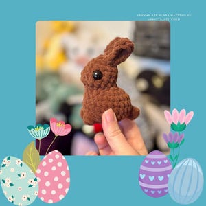 Peut inclure: Un petit lapin au crochet marron avec un œil noir. Le lapin est tenu dans une main et se détache sur un fond bleu avec des œufs de Pâques et des fleurs décoratives. Le texte en haut indique "CHOCOLATE BUNNY PATTERN BY @BRITTS_STITCHED."