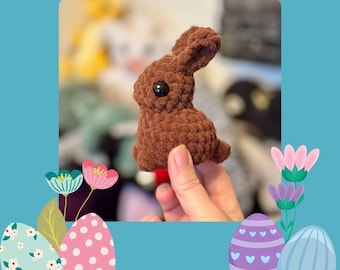 May include: A small, brown crocheted bunny with a black eye. The bunny is held in a hand and is set against a blue background with decorative Easter eggs and flowers. Text at the top reads "CHOCOLATE BUNNY PATTERN BY @BRITTS_STITCHED."