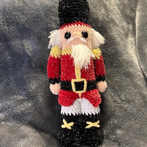 Könnte beinhalten: Handgemachte Nussknacker-Puppe aus Häkelarbeit. Die Puppe hat einen schwarzen Hut, eine rote Jacke mit goldenen Akzenten und eine schwarze Hose mit goldenen Kreuzen. Der Nussknacker hat einen weißen Bart und Schnurrbart sowie schwarze Augen.