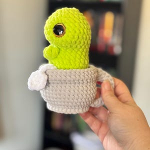 May include: A green crocheted plush toy with large eyes sits inside a gray crocheted teacup with a white handle.