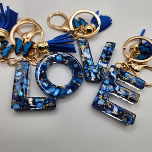 May include: Blue and gold resin keychain with the word "LOVE" spelled out in large letters. Each letter is decorated with gold flecks and blue and white iridescent pieces. The keychain has a blue tassel and a gold ring.