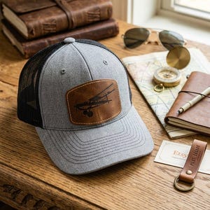 May include: A gray baseball cap with a black mesh back and a brown leather patch on the front. The patch features a dark brown embossed graphic of a small propeller airplane. The cap has a structured crown, a curved brim, and a button on top.