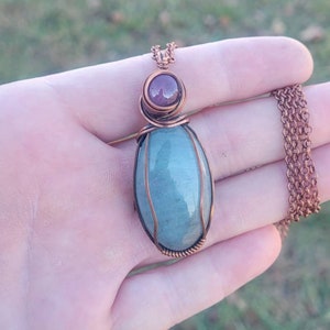 Op de afbeelding: Een hanger omwikkeld met koperdraad met een lichtblauwe ovale edelsteen en een kleinere, ronde, paarse edelsteen. De hanger hangt aan een koperen ketting.
