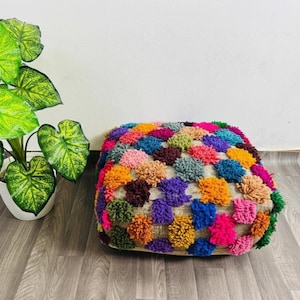 Op de afbeelding: Een vierkante, veelkleurige poef met een textuur. De poef is bedekt met een verscheidenheid aan kleurrijke pompons, waaronder roze, oranje, blauw, groen en paars. De basis lijkt van natuurlijk jute materiaal te zijn.