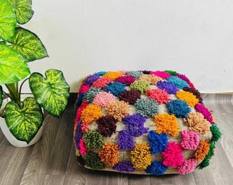 May include: A square, multi-colored pouf with a textured surface. The pouf is covered in a variety of colorful pom-poms, including pink, orange, blue, green, and purple. The pouf is placed on a wooden floor next to a potted plant.