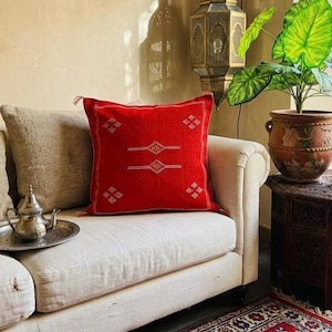 May include: A red square throw pillow with white and gold geometric designs rests on a white sofa. The pillow has a textured appearance and is adorned with tassels. A silver tea set sits on a tray on the sofa.