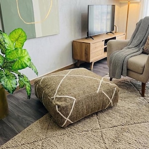 Peut inclure: Un pouf carré marron avec un motif géométrique blanc est posé sur un tapis texturé. Le pouf est dans un salon, avec un meuble TV en bois, un fauteuil gris et une plante verte en arrière-plan.
