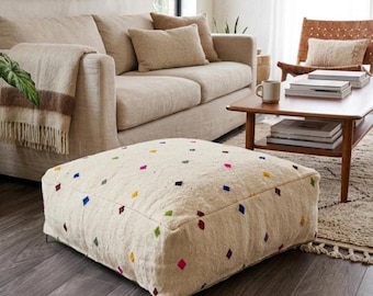 May include: A square, off-white pouf with colorful diamond-shaped accents. The pouf is placed on a dark wood floor in a living room setting. A beige sofa and coffee table are visible in the background, creating a cozy atmosphere. The pouf is a decorative and functional piece of furniture.