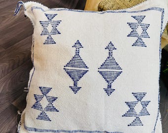 May include: Square white cushion with a geometric blue pattern. The cushion has a textured weave and blue fringe on the corners. The design features diamond and triangle shapes. Decorative cushion for a sofa or bed.