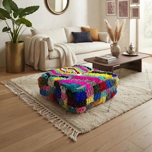 May include: A square floor pouf featuring a thick, shaggy, multi-colored pile with a vibrant geometric pattern in shades of hot pink, bright blue, yellow, green, and deep red, resting on a textured cream-colored area rug with fringe edges.
