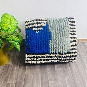 May include: A square, textured floor cushion with a geometric design. The cushion features a vibrant blue square, a gray rectangle, and black and white striped borders. The plush material adds a cozy, decorative element to any room.