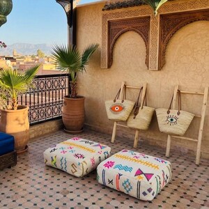 Peut inclure: Deux coussins de sol carrés en tissu blanc avec des motifs géométriques colorés, trois sacs fourre-tout en paille tressée avec des anses en cuir et des motifs d'yeux brodés suspendus à des échelles en bois, et deux palmiers en pot.