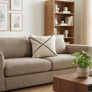 Peut inclure: Un canapé beige texturé avec un coussin carré blanc cassé orné d'un motif en X cousu noir. Le canapé présente une finition en tissu bouclé et est placé dans une pièce avec une bibliothèque en bois et une plante verte sur une table en bois.