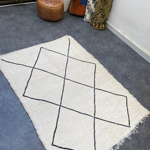 May include: An off-white rectangular rug featuring a black geometric diamond design. The rug has a fringed edge and is shown on a dark grey surface. Rolled rugs and a leather ottoman are visible in the background, adding to the decor.