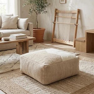 Op de afbeelding: Een neutraal getinte woonkamerscène met een beige vierkante poef met een geometrisch ontwerp. Een houten salontafel, een bank en een vloerkleed vullen de poef aan. Een weefgetouw en een olijfboom in pot dragen bij aan de inrichting van de kamer.