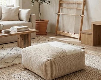 May include: A neutral-toned living room scene featuring a beige square ottoman with a geometric design. A wooden coffee table, sofa, and rug complement the ottoman. A weaving loom and potted olive tree add to the room's decor.