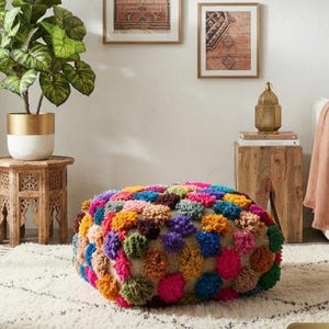 May include: A square floor pouf featuring a dense, textured surface covered in multi-coloured yarn pom-poms in shades of pink, blue, yellow, green, and purple, arranged in a grid pattern over a neutral-toned woven fabric base.