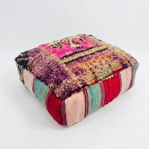 May include: A square floor pouf featuring a thick, textured, hand-knotted wool top in vibrant shades of magenta, purple, orange, and green. The sides are constructed from flat-woven fabric with vertical stripes in red, teal, and cream colours.