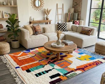 May include: A colorful, geometric patterned rug with a white fringe sits on a dark wood floor. The rug features orange, blue, green, and black shapes. A beige sectional sofa and wooden coffee table are in the background.