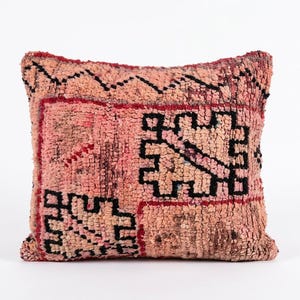 Peut inclure: Un coussin carré avec un poil noué à la main texturé dans des tons de rose poudré et saumon. La surface est décorée de motifs géométriques noirs audacieux et de fines lignes rouges formant des formes rectangulaires et abstraites.
