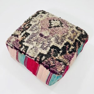 Peut inclure: Un coussin de sol carré dont la surface supérieure présente un poil épais et touffu dans des tons crème, anthracite et violet sourd avec un motif géométrique. Les côtés sont en tissu lisse avec des rayures verticales roses, rouges et bleu sarcelle.