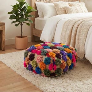 Op de afbeelding: Een vierkante vloerpoef met een dicht, getextureerd oppervlak bedekt met veelkleurige garen pompons in tinten roze, oranje, geel, groen, blauw en paars, geplaatst op een neutrale beige geweven basisstof.