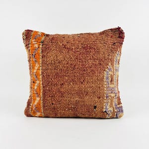 Peut inclure: Un coussin carré avec un panneau central texturé brun rouille. Le côté gauche présente une bande verticale orange avec un motif en zigzag, tandis que le côté droit affiche un motif géométrique violet clair et crème sur fond sombre.
