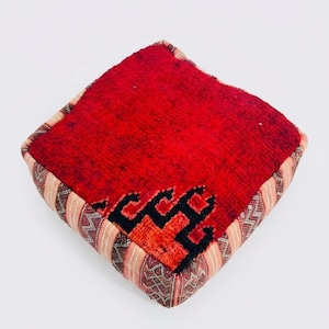 May include: A square floor cushion featuring a vibrant red textured top surface with a black geometric pattern near one corner. The sides are constructed from a striped fabric in shades of beige, brown, and red with intricate woven detailing.